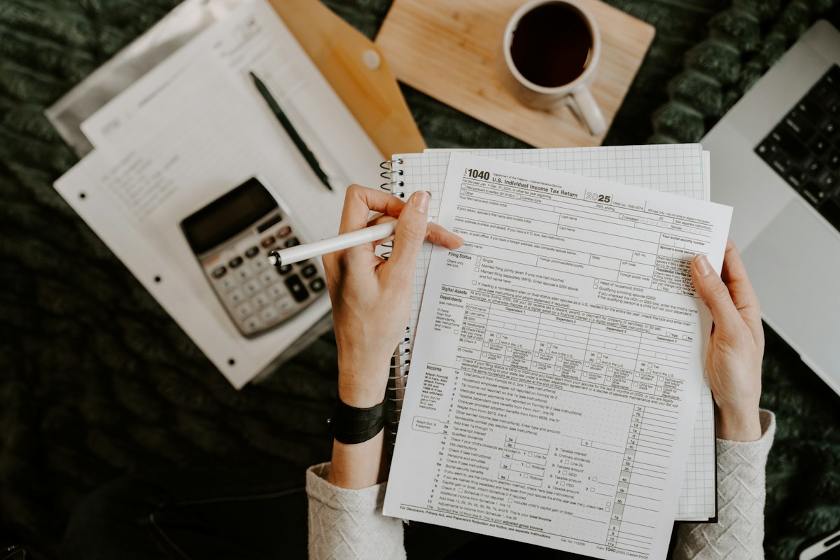 Overhead view of hands holding a pen over a U.S. 1040 tax form, with a calculator, notebook, coffee mug and laptop on the table