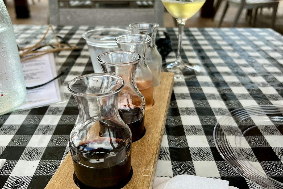 Wine flight of four small glass carafes holding red, rosé and white wines on a wooden paddle, on a black-and-white checked tablecloth with a glass of white wine behind